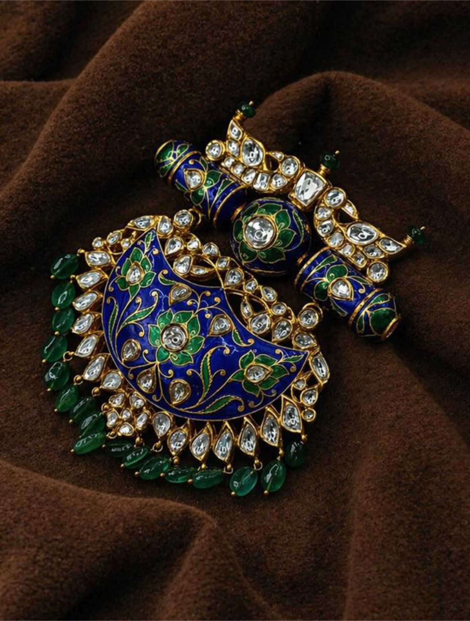 Pair of gold peacock pendant earrings with blue enamel, floral details, diamonds, and green beads on brown fabric.