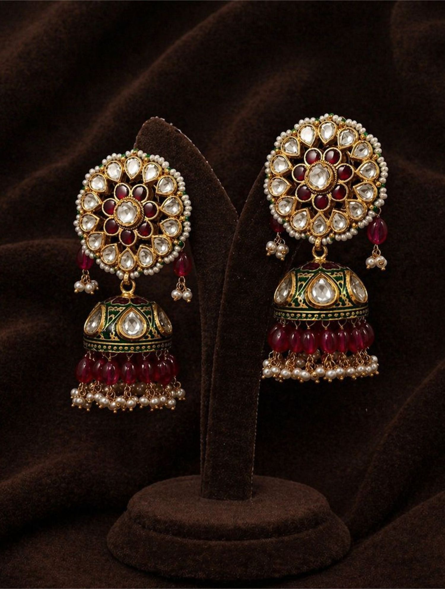 Pair of gold kundan jhumka earrings with ruby stones, pearls, and green enamel bells on velvet stand.