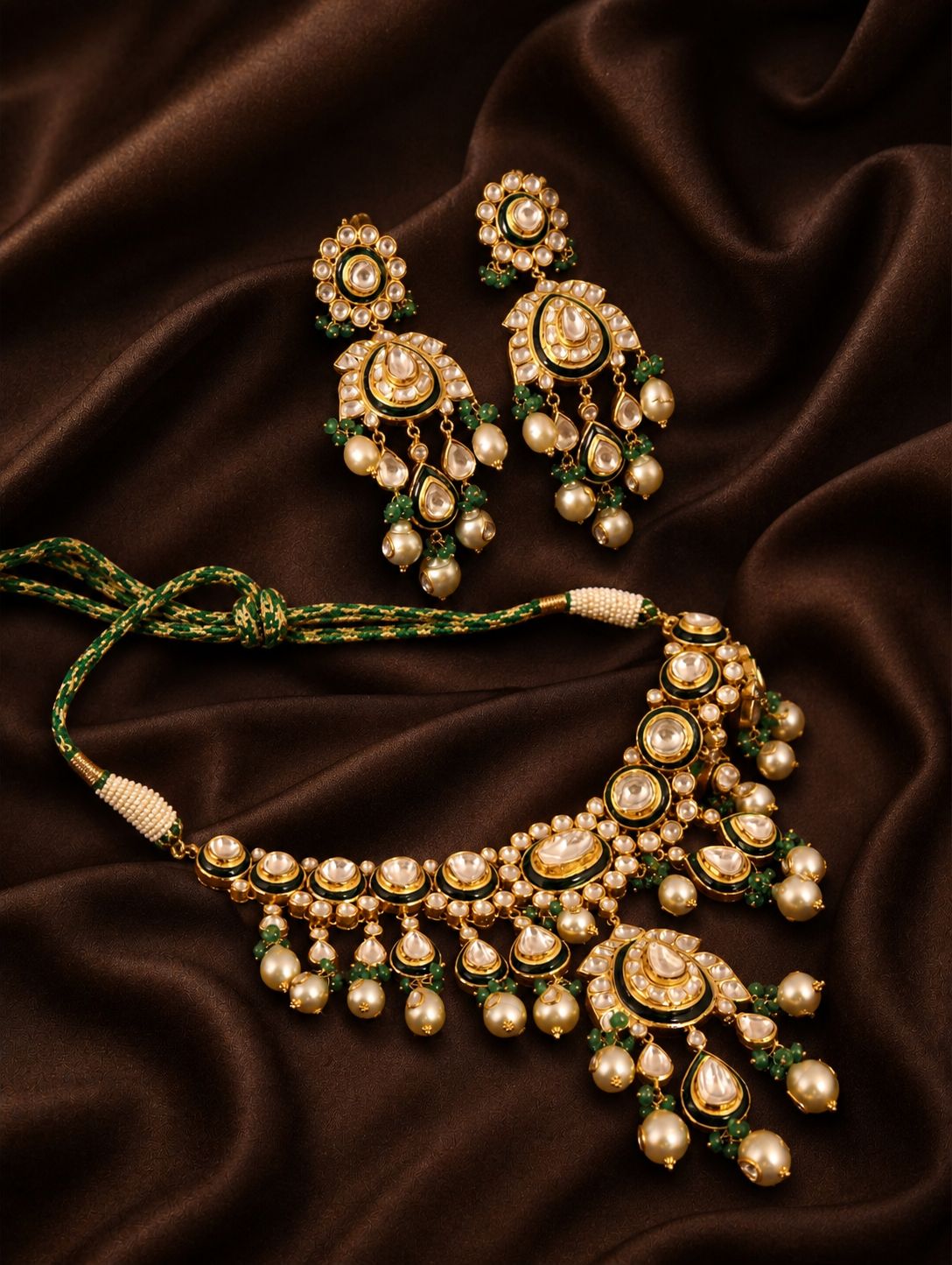 Gold kundan jewelry set with earrings and necklace featuring crystals, pearls, and green stones on brown silk fabric